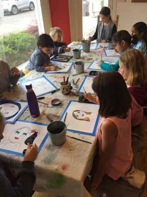 A class of young students sitting around a table using watercolors to paint in their line drawing self portraits